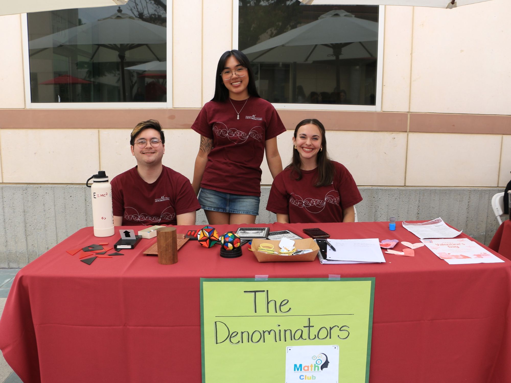 Three students stand behind a table labeled “The Denominators," displaying puzzles and informational materials. 