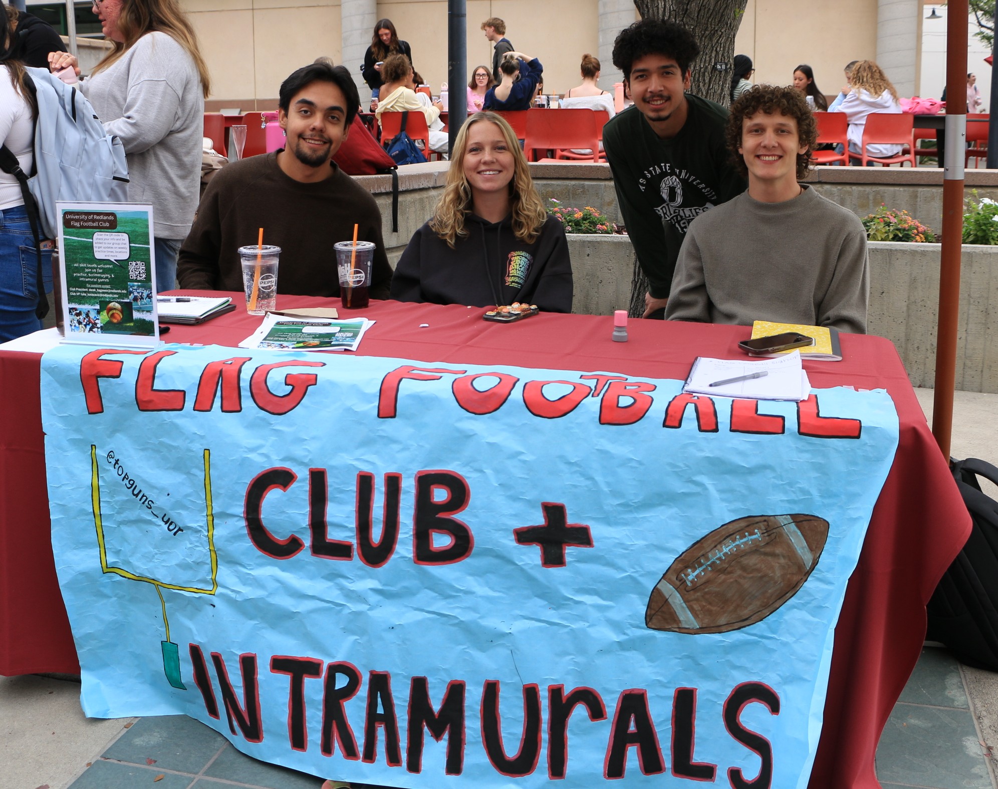 Four students smile while gathered behind a table with a banner for Flag Football Club and Intramurals.