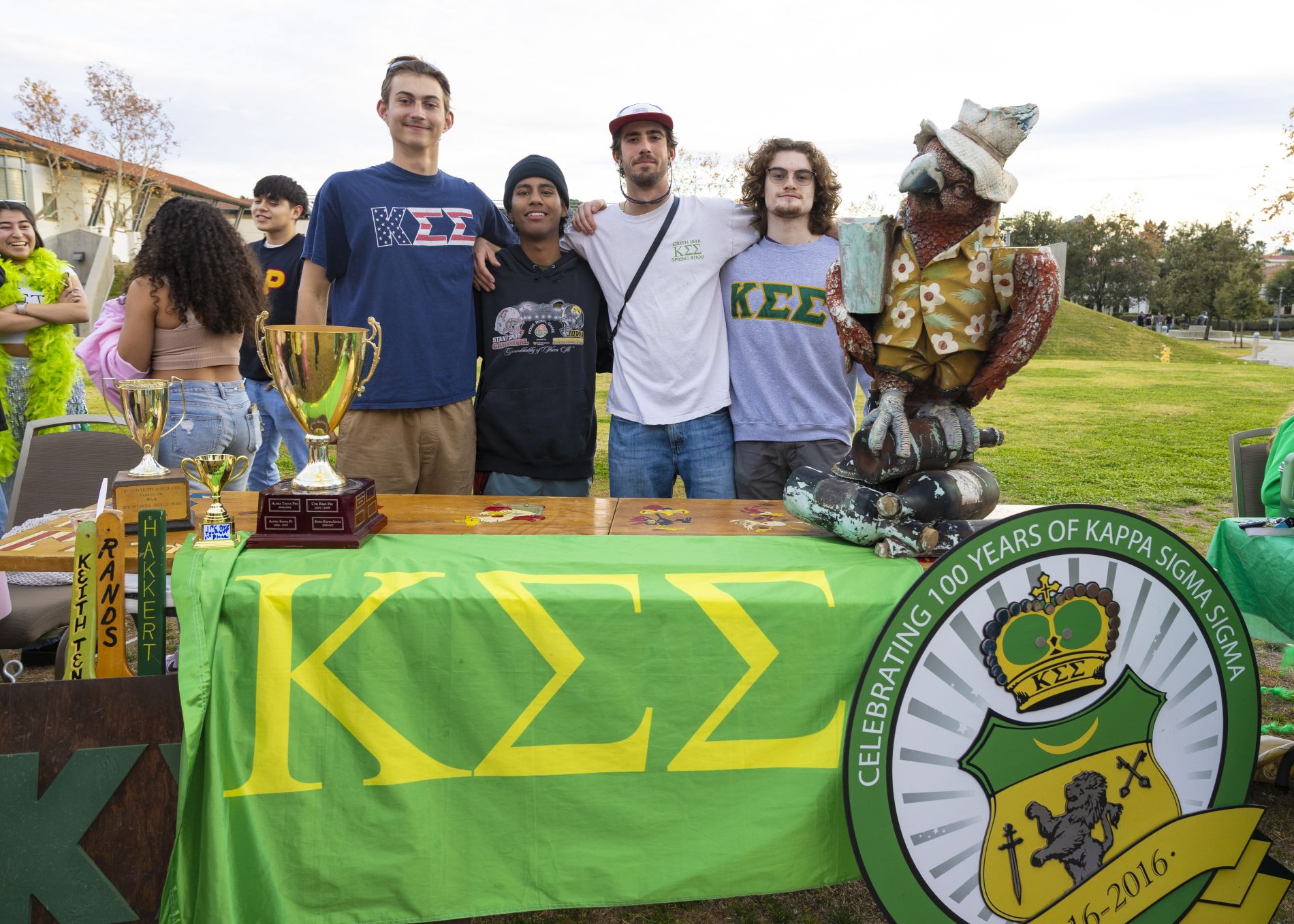 Five students stand behind a table displaying trophies, memorabilia, a green Kappa Sigma Sigma banner, and a decorative statue at a outdoor campus involvement fair.